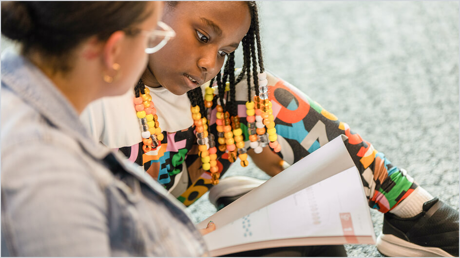 Two middle school students sit together on the floor, discussing a page in a math workbook.
