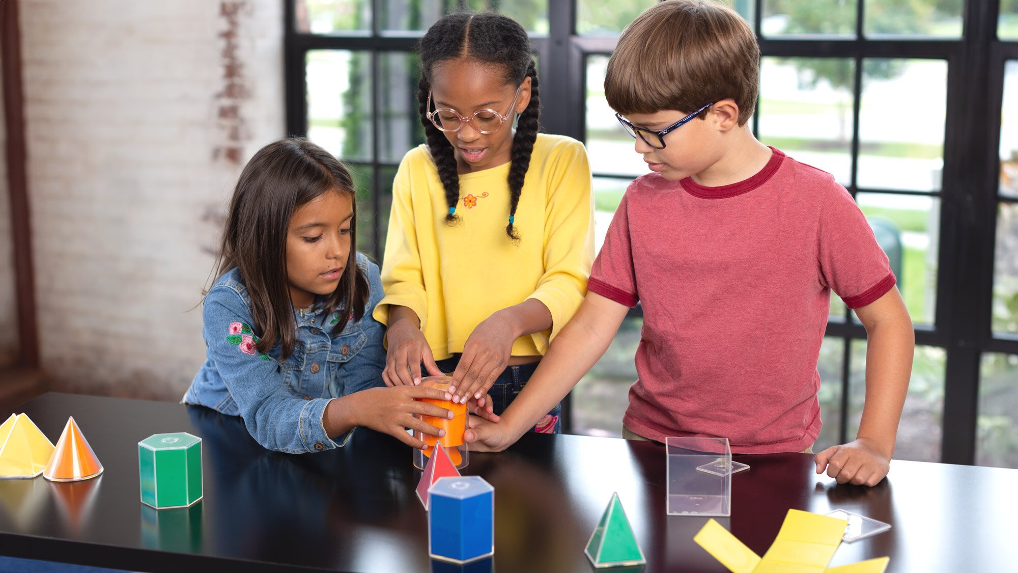 Three elementary students working with 3D shapes at a classroom table. Three elementary students working with 3D shapes at a classroom table.