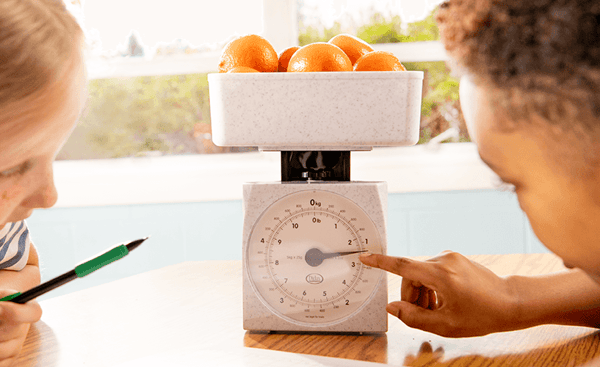 Two children examine a kitchen scale holding several oranges. One child points to the dial while the other takes notes with a pencil, suggesting a math or measurement activity. Two children examine a kitchen scale holding several oranges. One child points to the dial while the other takes notes with a pencil, suggesting a math or measurement activity.