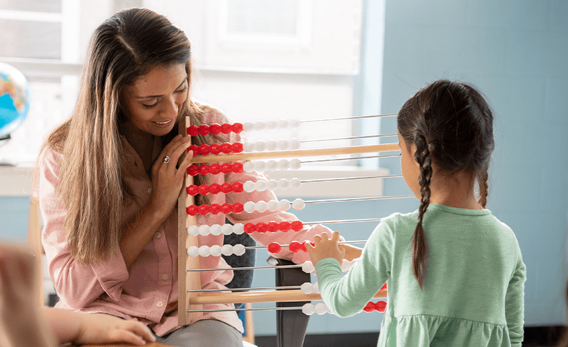 A teacher sits beside a student who is moving beads on a large red-and-white rekenrek during a math activity in a classroom.