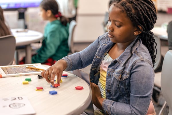 A student arranges colorful rectangular tiles on a classroom table while working through a Eureka Math Squared activity, with a classmate visible in the background.