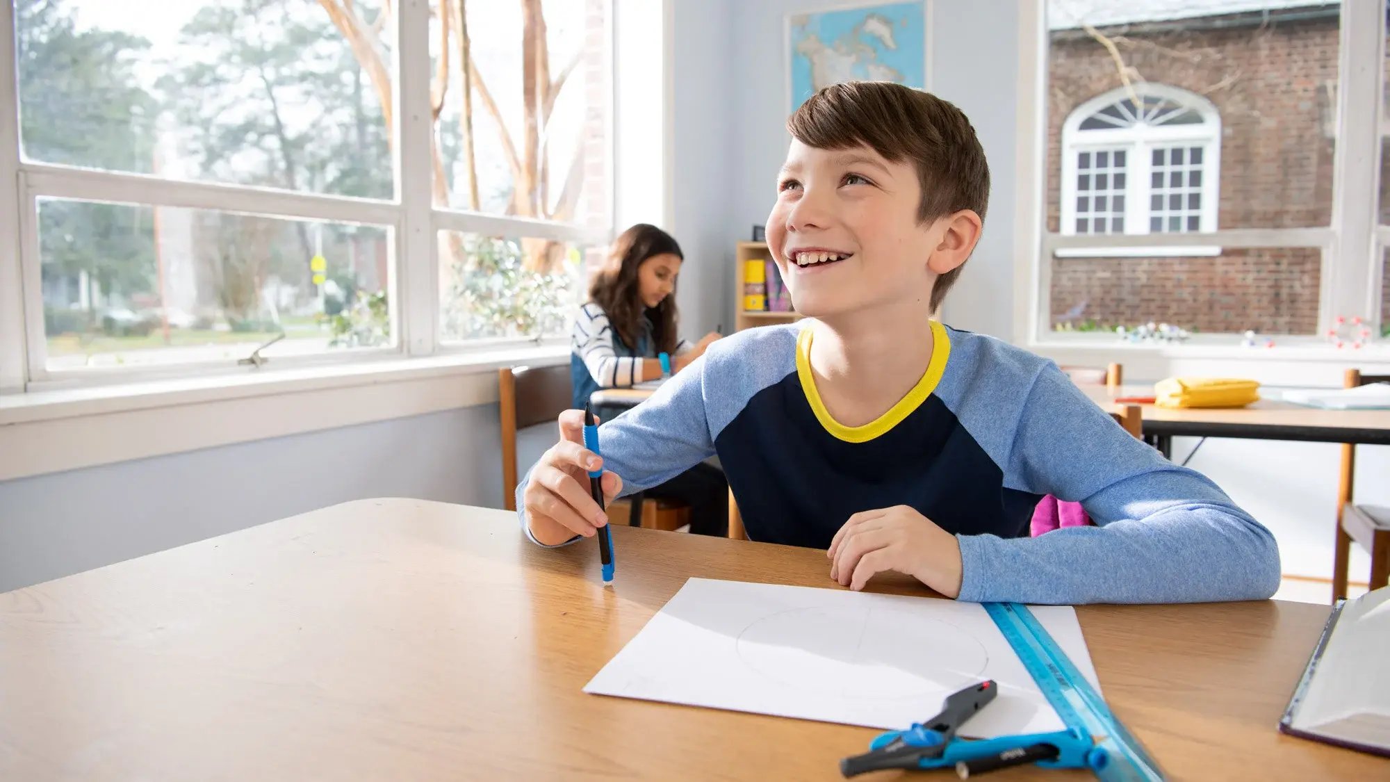 Male elementary student seated at classroom table holding a mechanical pencil with a female elementary student seated at another classroom table in the background.