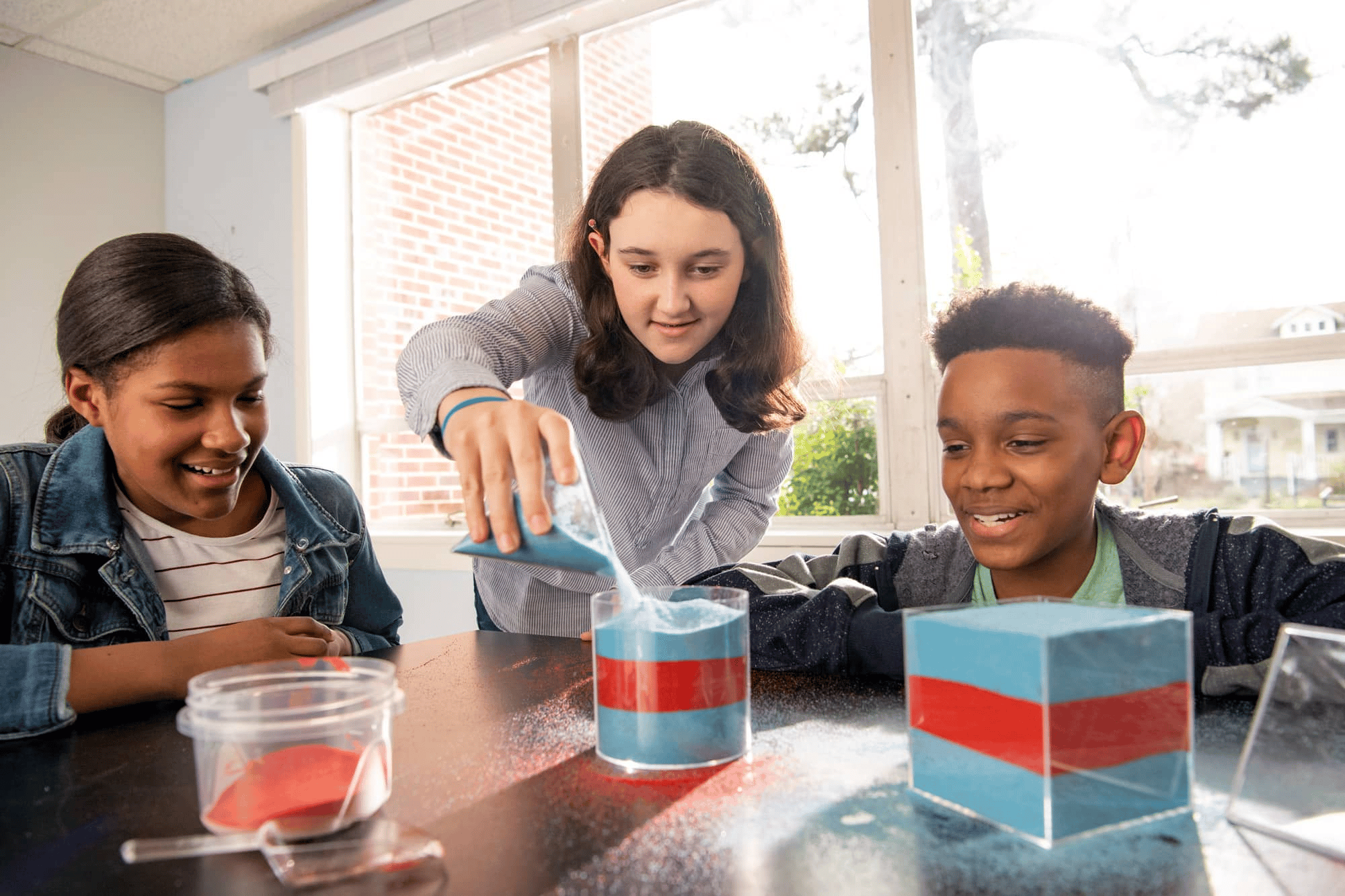 Three middle school students working with transparent containers and sand at a classroom table.