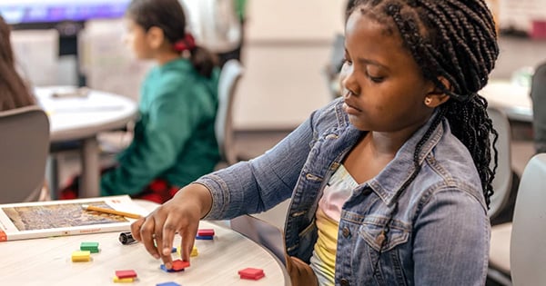 A student arranges colorful rectangular tiles on a classroom table while working through an Eureka Math Squared activity, with a classmate visible in the background.