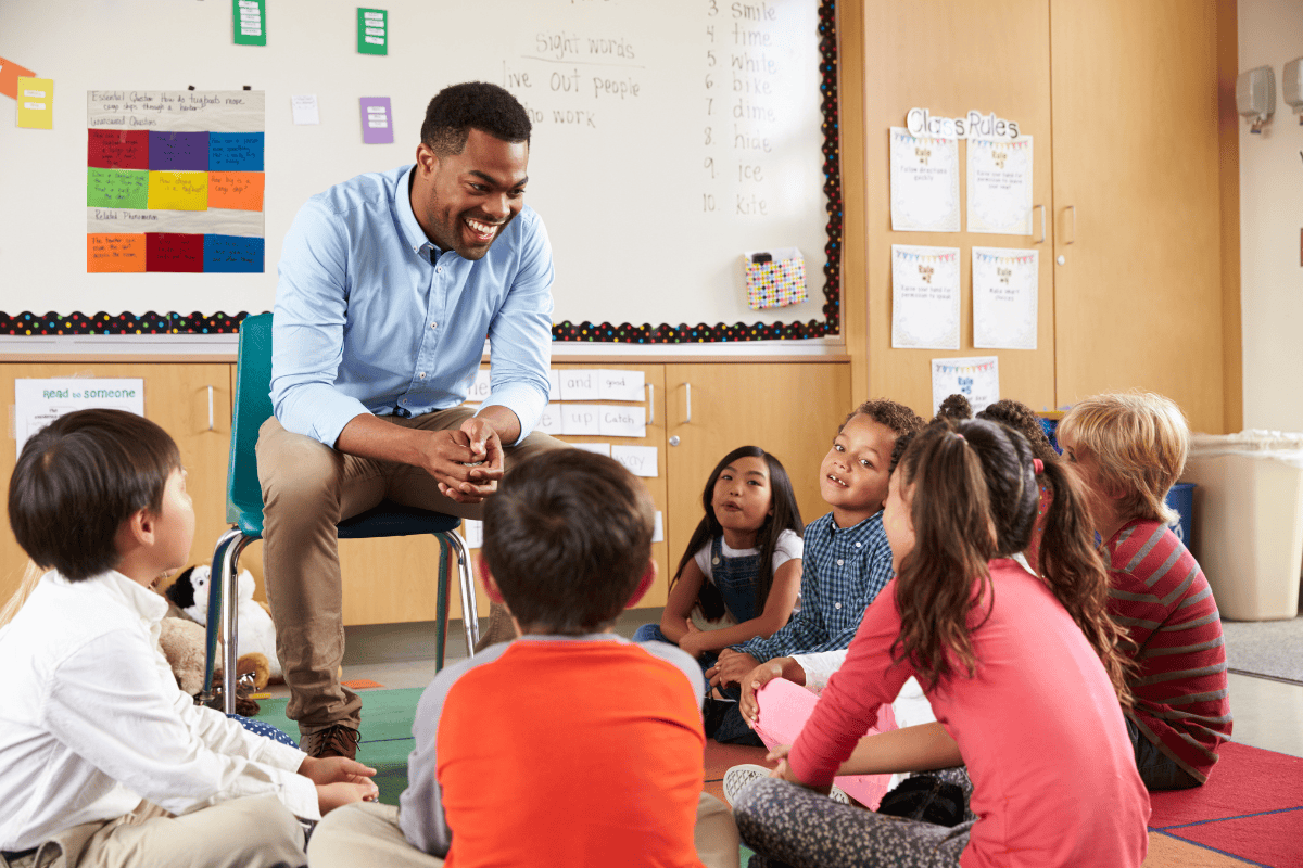 Teacher speaking with his students while they are seated on the floor of the classroom.