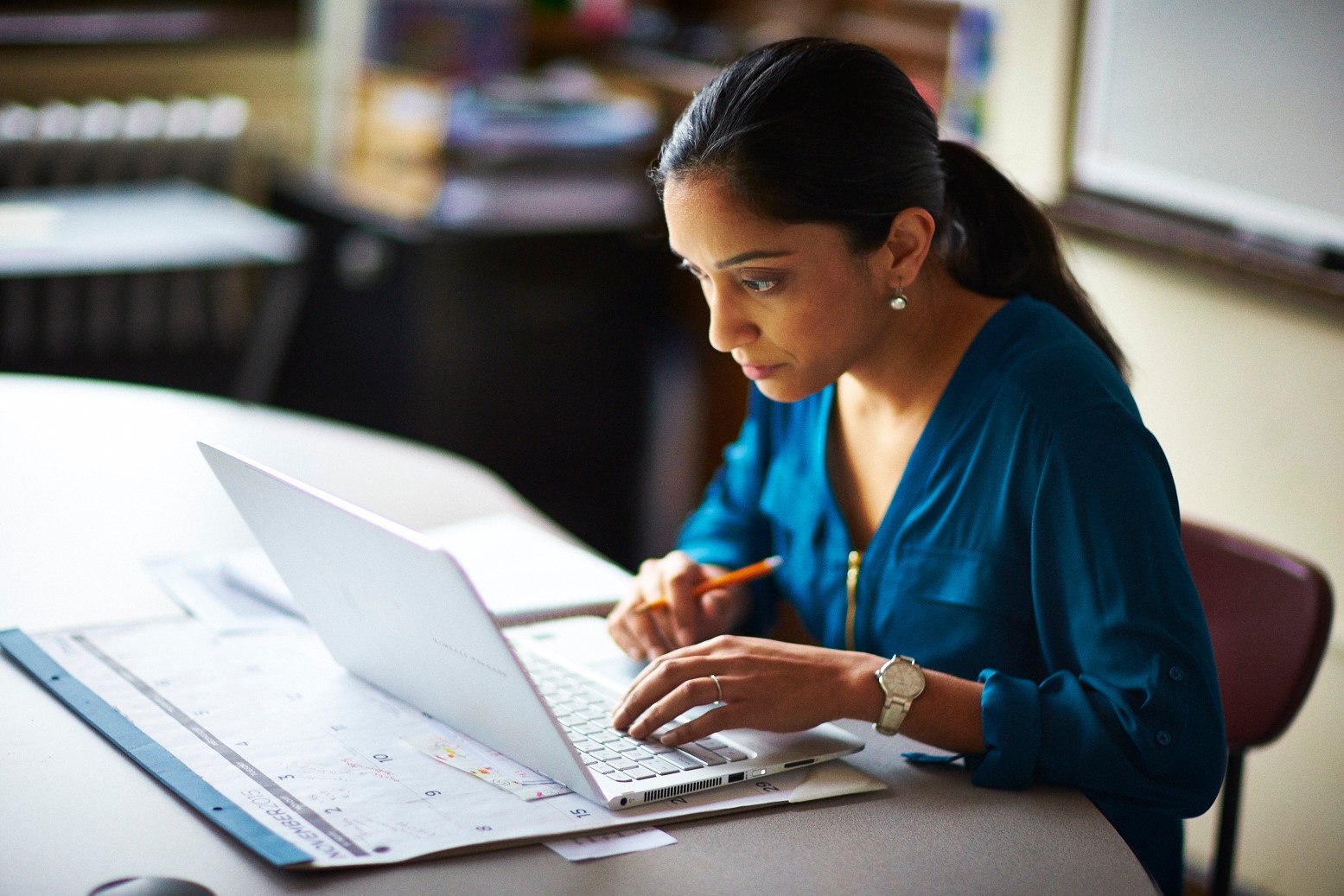 A teacher working at her desk in her classroom on a laptop.
