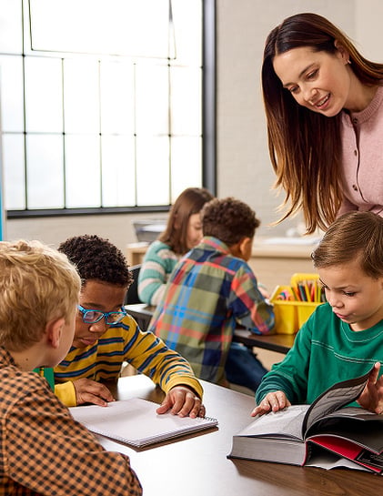 A teacher leans over a table to help young students as they read and write together in a classroom. A teacher leans over a table to help young students as they read and write together in a classroom.