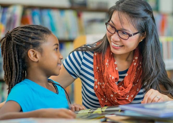 Female elementary student sitting in a classroom at a desk working with her teacher.