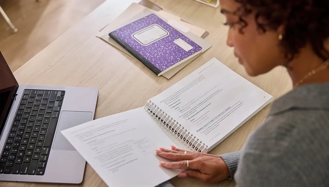 A person reviewing a spiral-bound curriculum sampler and printed pages at a desk with a laptop