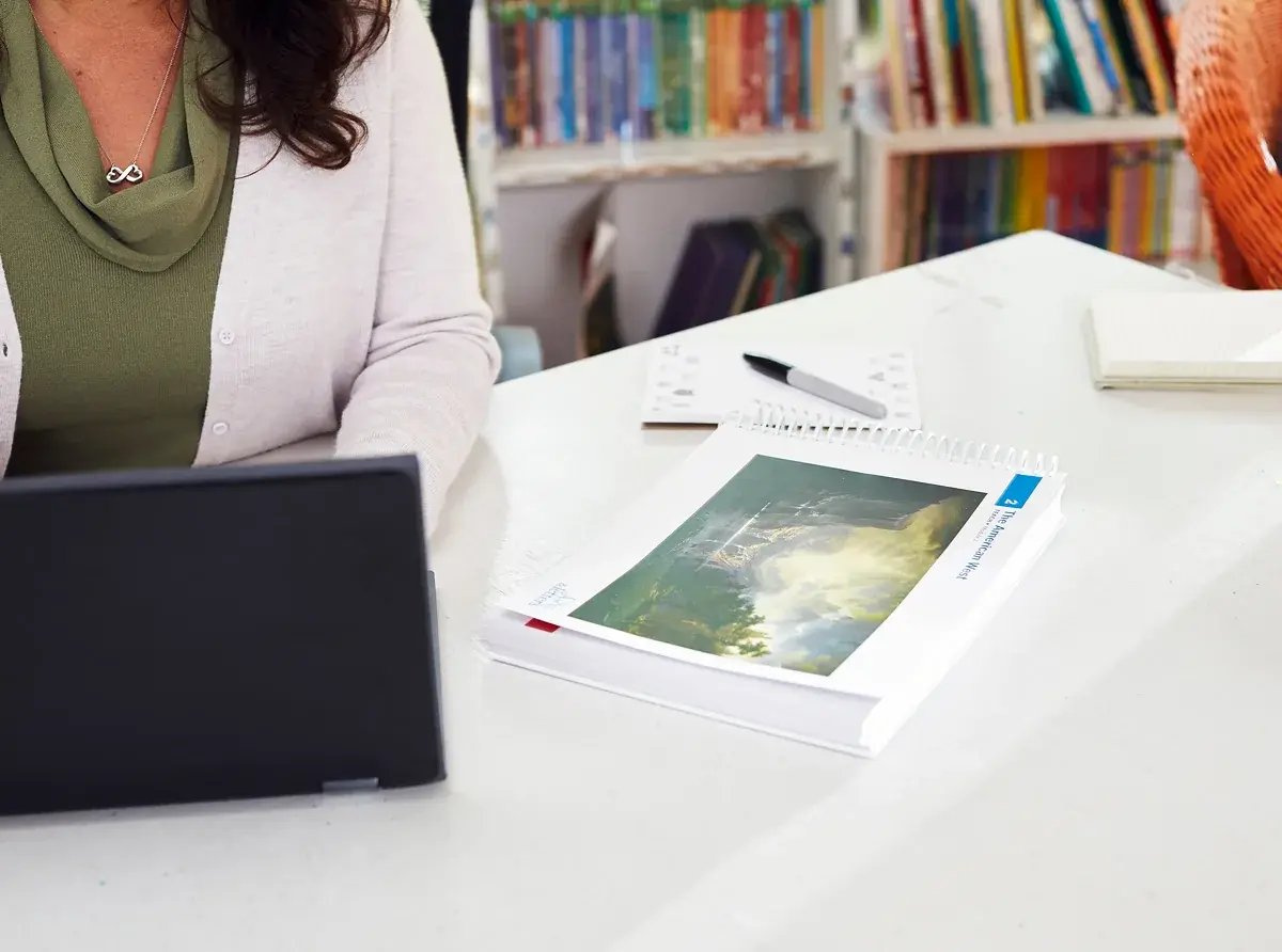 A teacher at a laptop beside an open Arts & Letters guide on a classroom table, with shelves of books in the background