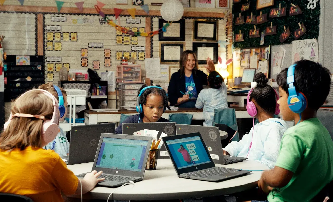 Young students wearing headphones and working on laptops at a table while a teacher leads the class in a decorated classroom
