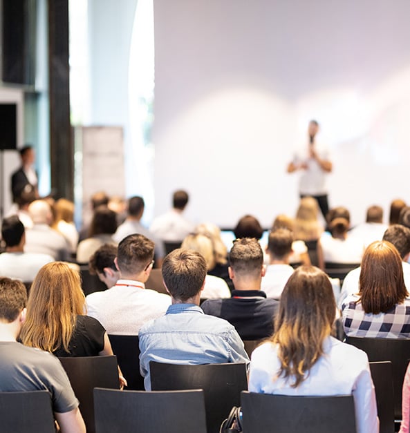 A large audience sits facing a speaker presenting at the front of a conference or seminar room. A large audience sits facing a speaker presenting at the front of a conference or seminar room.