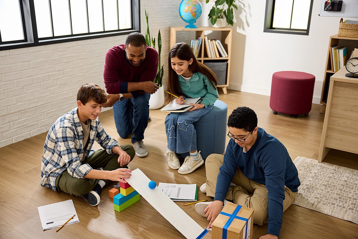 A teacher kneels on the classroom floor with three students as they conduct a hands-on activity. The students sit around a ramp made from a board and use colorful blocks, a ball, and a box to experiment, with notebooks and pencils nearby.